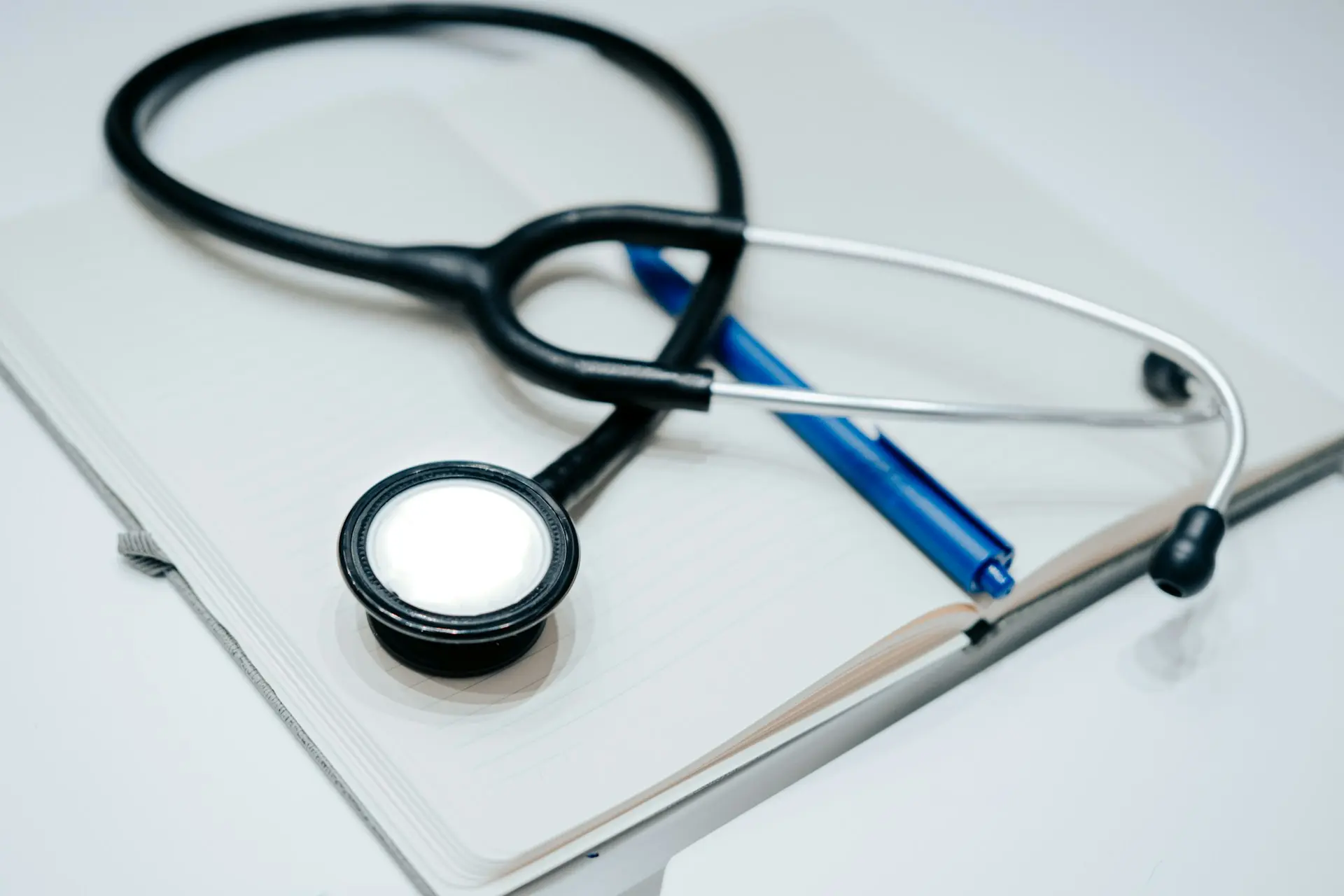 A black stethoscope and blue pen resting on an open notebook against a white surface.
