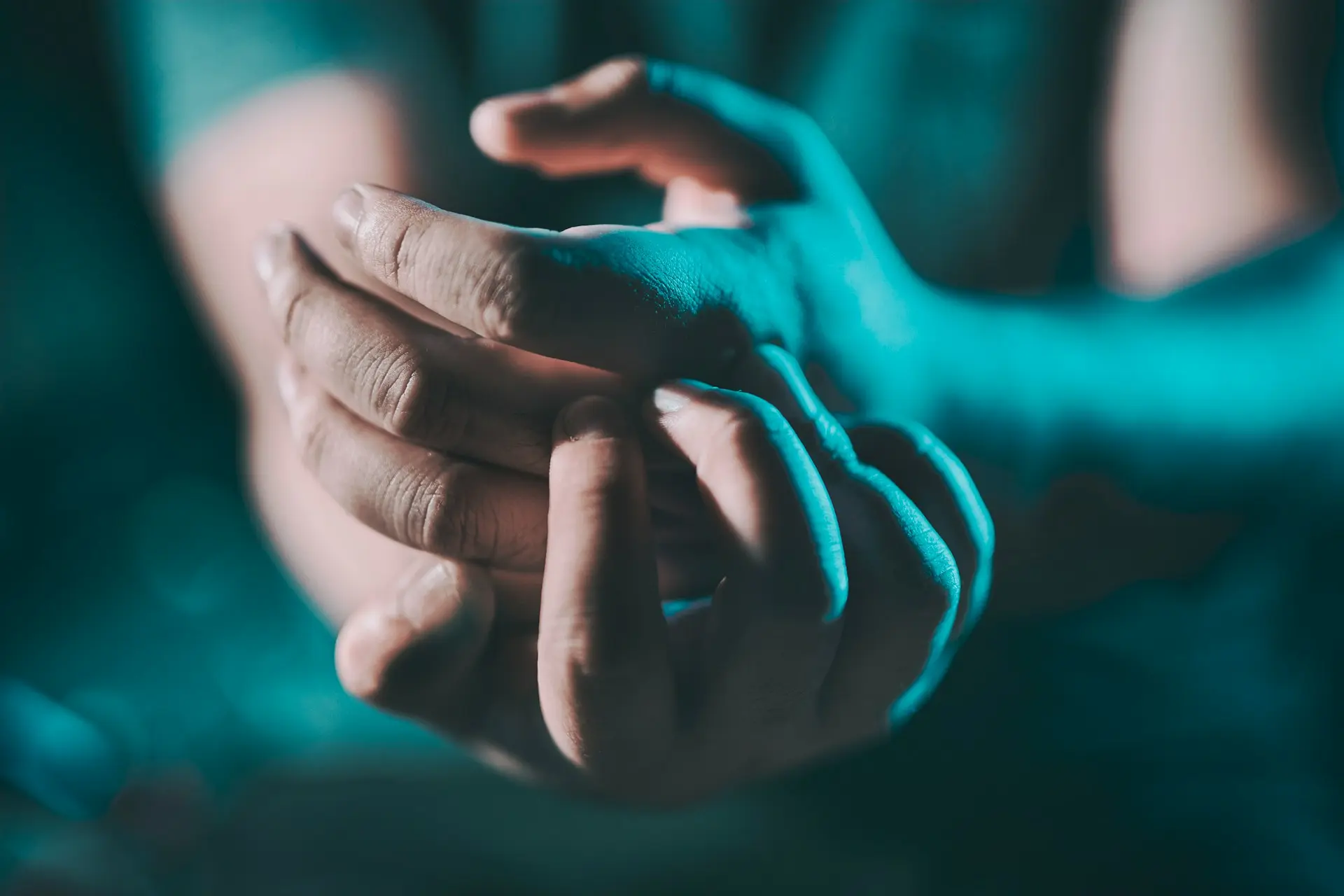 Close-up of a person’s hands under teal lighting, gently holding and rubbing one finger with the other hand, suggesting discomfort or joint pain.
