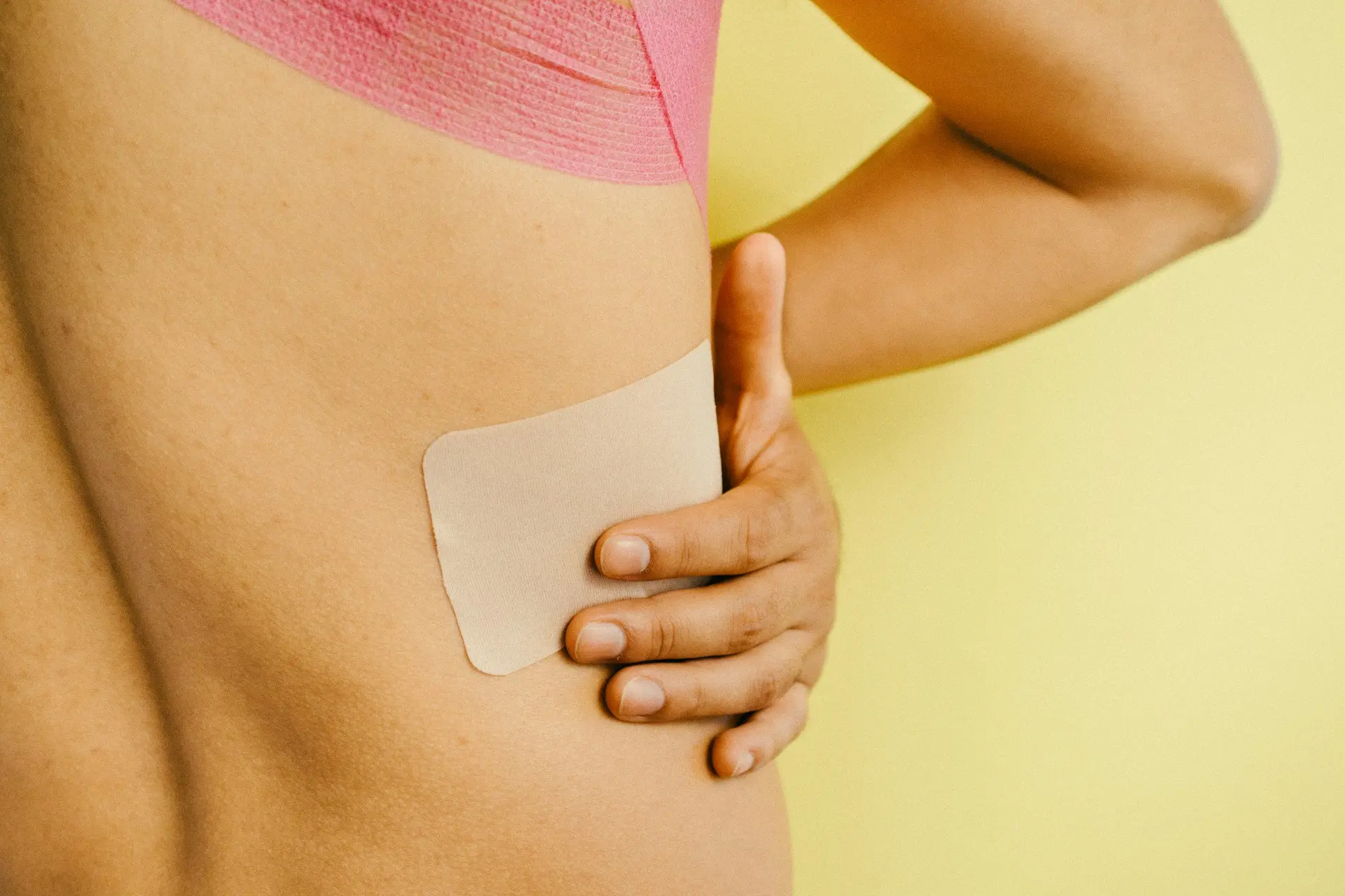 A close-up of a person pressing a beige transdermal patch onto the side of their abdomen against a yellow background.